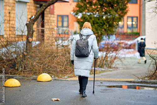 Woman with cane walking through wet residential courtyard where melting snow, puddles, and winter remnants create slippery environment, mobility challenges. Woman walk with help of cane