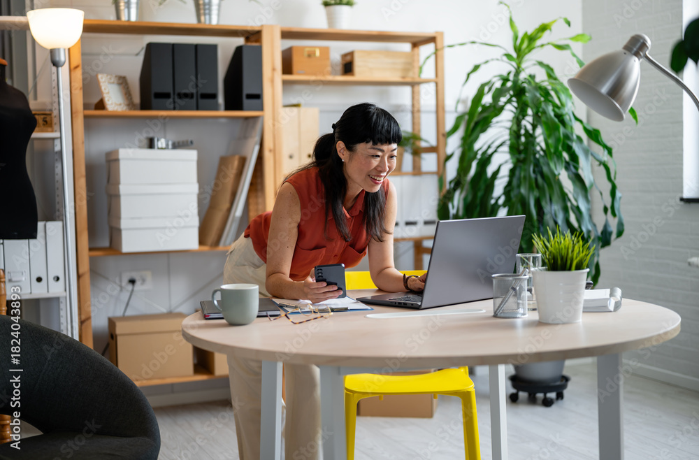 Fototapeta premium Asian woman small entrepreneur working in her cozy home office, standing at her desk while reviewing notes and using her laptop in a bright, modern workspace