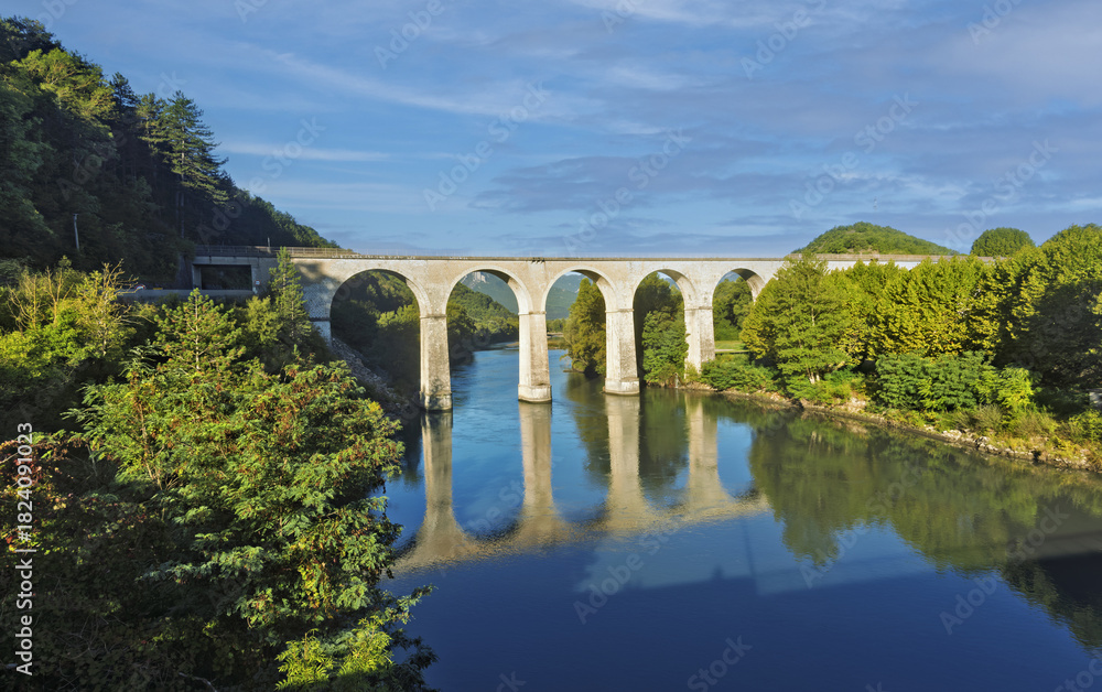 Fototapeta premium Stone arch bridge in Sisteron, France, with multiple evenly spaced arches reflected in the water. the bridge stands out for its symmetry and architectural elegance under clear daylight. 