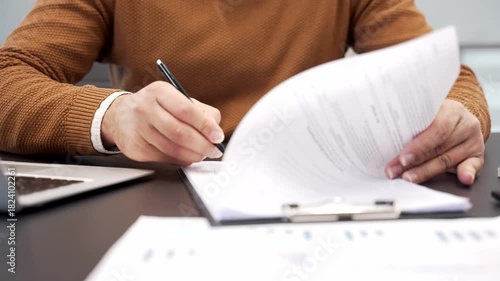 Close up of a businessman's hand signing documents at a desk at a workplace in a business office. An executive or manager in casual sweater reviews the documentation and signs the contract with a pen