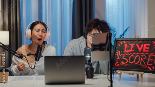 Young happy Asian couple wear headphone sit on desk online live streaming football live score looking at camera watch soccer sport game on laptop talk with follower in small studio at home at night.
