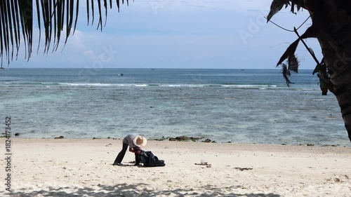 A person in a straw hat cleans up trash on a pristine tropical beach, framed by palm leaves, emphasizing environmental protection and pollution.