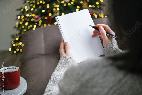 Over-the-shoulder view of person in sweater writing in blank spiral notebook with pen with blurry Christmas tree and lights in background. Goals, wish list, resolutions, planning, to-do list, mockup