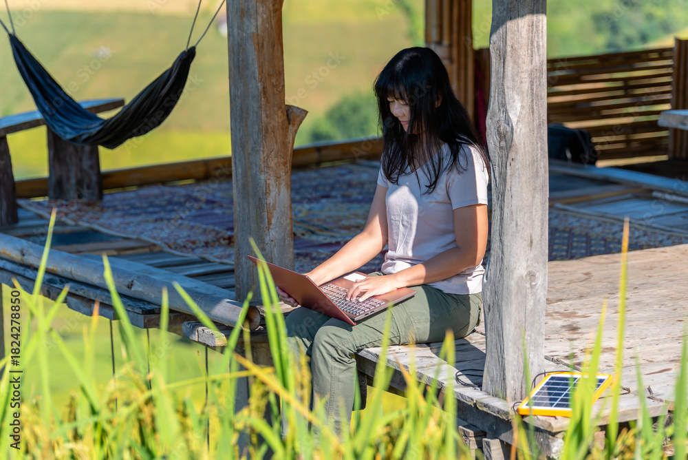Obraz premium Asian woman working on a laptop outdoors using portable solar panel for sustainable charging. Remote work, digital nomad, and eco-friendly technology concept.