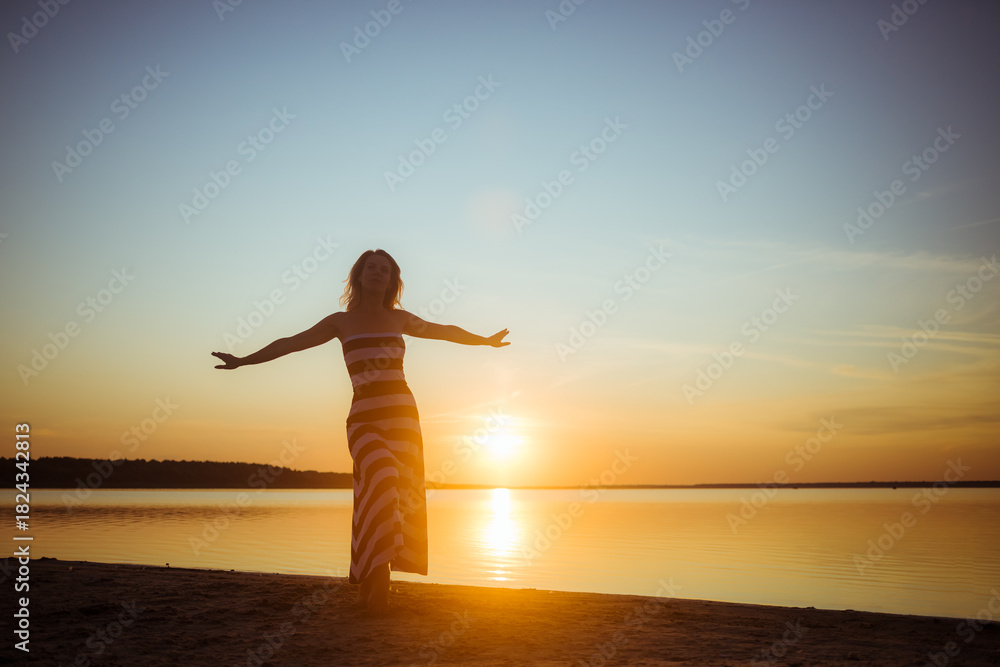 Fototapeta premium Silhouette of young sexy woman in full-length stands with an arms are outstretched against the sunset on the lake's shore in long striped dress. Greeting to the sun. Women's freedom. Summer vibe.