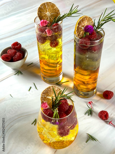 High-angle view of three refreshing golden cocktails garnished with fresh raspberries, dried lemon slices, rosemary, and edible flowers served on a white marble table with loose ingredients nearby.