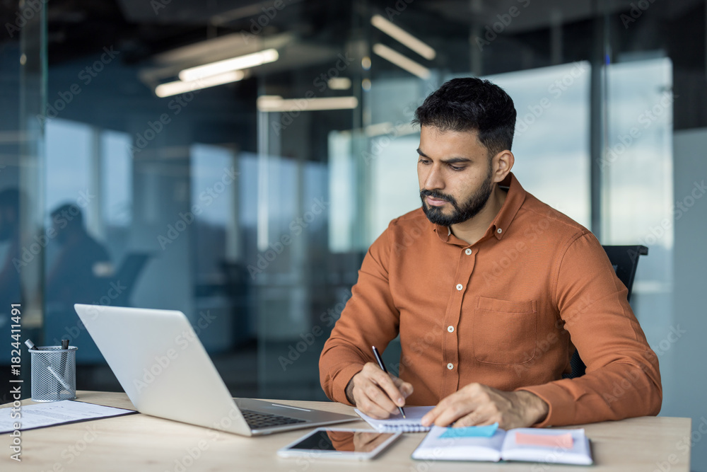 Obraz premium Young businessman concentrating on taking notes in a notebook at his desk, having a laptop and tablet, during a productive workday in a contemporary office environment