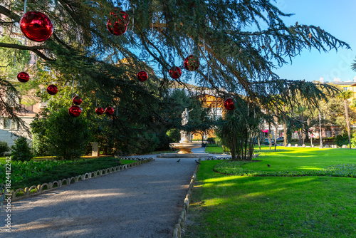 Large red Christmas ornaments hanging from tree branches in a sunny city park in Opatija with a fountain in the background