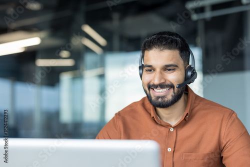 Young indian man in smart casual wearing a headset, smiling while providing online customer support at a laptop in a modern office workspace, professional and approachable during a video call