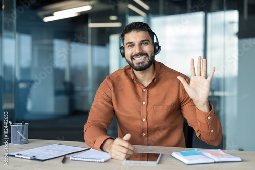 Young indian man wearing a headset and an orange shirt, smiling and waving, communicating online from a modern office environment during a virtual meeting
