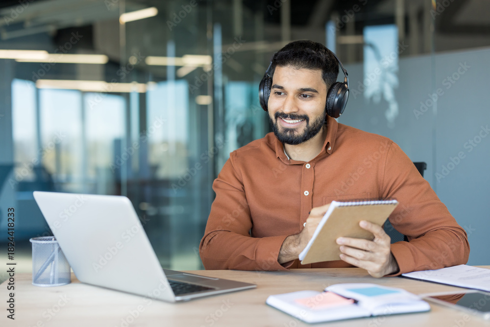 Fototapeta premium Smiling young man in an open office setting wearing headphones, actively participating in a virtual meeting or online course on a laptop while taking notes in a notebook