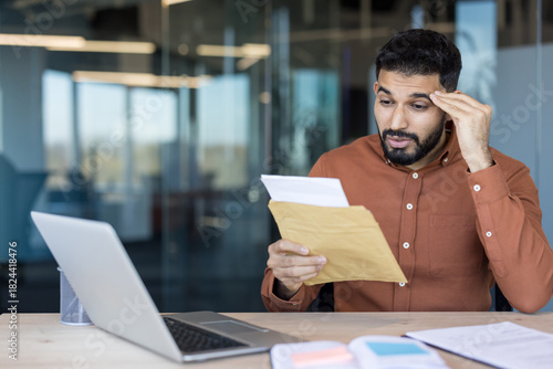 Stressed young man discovering shocking information in a letter, feeling worried and surprised while sitting at his desk in a modern office environment