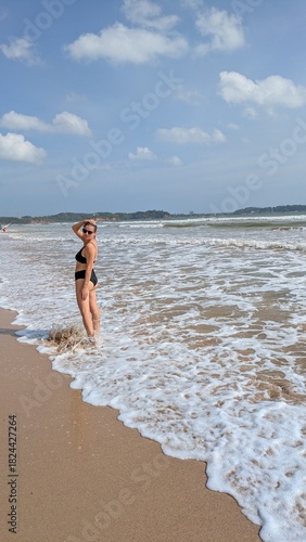Tanned sensual woman smiling and laughing, enjoying a tropical vacation escape in paradise, walking barefooted in bikini swimwear along the ocean wave sand shore with palm, eyewear and sunglasses