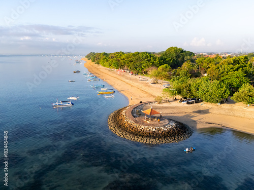 Fototapeta Naklejka Na Ścianę i Meble -  Sunny coastal landscape with calm blue waters, traditional boats, and a stone pavilion overlooking a peaceful sandy beach lined with lush green trees. Perfect serene tropical scenery.