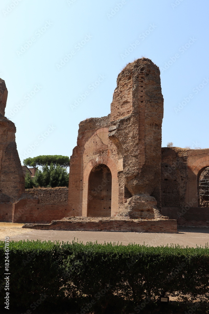 Naklejka premium A dramatic, tall section of ancient Roman bath ruins, featuring a massive brick archway framed by green shrubs