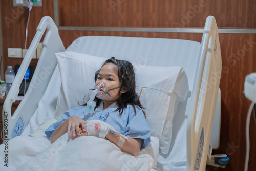 Sick Asian girl wearing oxygen mask lying on hospital bed receiving treatment. Healthcare concept, respiratory disease, hospital admission, medical care, and child patient in emergency situation.