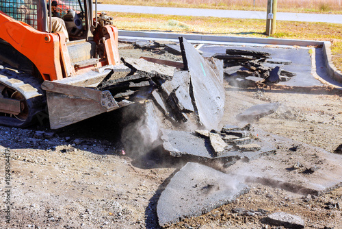 Construction skid steer machinery is breaking up asphalt in parking lot while dust rises into air