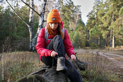 A young female hiker is lacing her trekking boots while sitting on a log in the forest. Hiking in the forest in autumn