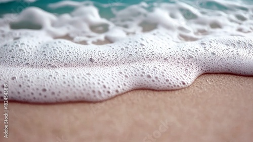 Fototapeta Naklejka Na Ścianę i Meble -  Close-up of gentle ocean wave foam washing onto a sandy beach, with soft turquoise water in the background. Natural daylight.