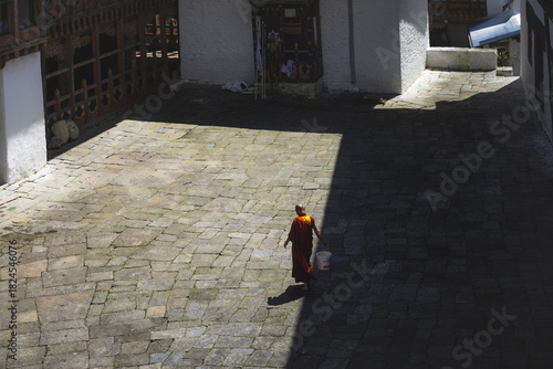 View of a solitary monk in vibrant orange robes walks across a sun-drenched stone courtyard, casting a long shadow in Trongsa Dzong, Trongsa, Bhutan.