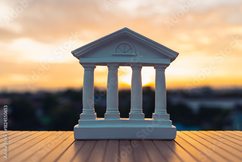 White model of classical bank building with pillars on wood table at sunset, symbolizing financial institutions, government stability, law, economy, investment security, and banking systems.