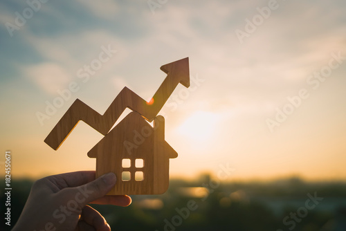 Fototapeta Naklejka Na Ścianę i Meble -  Hand holding wooden house with rising arrow graph at sunset, symbolizing real estate investment growth, property market trends, mortgage rates and housing price increase in financial concept.