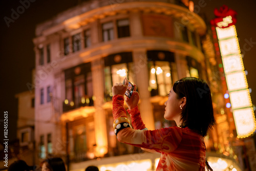 Woman in vibrant clothes photographs illuminated building at night in bustling city street. Shanghai, China