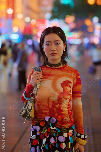 Young woman in a colorful outfit stands on a vibrant, busy city street at night. Shanghai, China