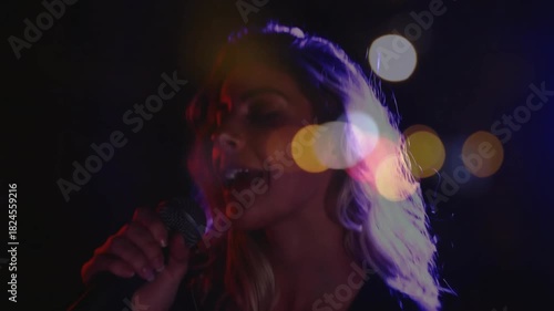 Female singer raising mic and singing, performing music on dim stage, bokeh lights crossing face