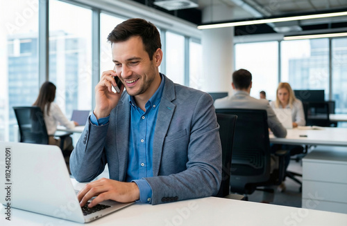 Smiling businessman multitasking with smartphone and laptop in a modern bright office with colleagues in the background