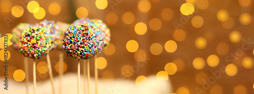 Colorful cake pops with bright sprinkles are displayed on sticks, adding joy to a festive holiday gathering. The warm lights create a cheerful atmosphere for Christmas and New Year 2026