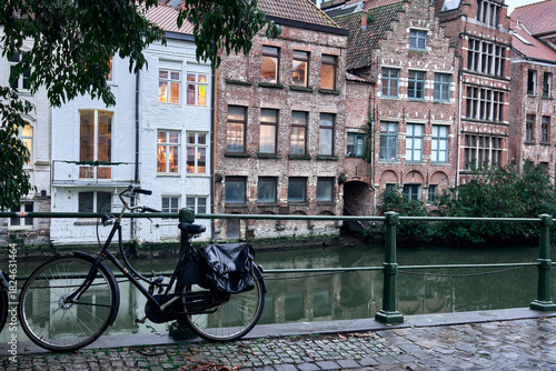 A scenic view of traditional Flemish buildings along the River Lys in Ghent, Belgium. A classic bicycle is parked by a green railing, surrounded by cobblestone and calm waters, under leafy branches. 