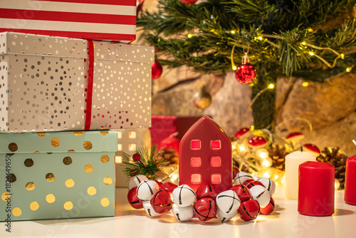 A Christmas setting: wrapped gift boxes, a glowing red ceramic house surrounded by jingle bells. A decorated Christmas tree with red baubles and warm string lights, pinecones and candles.