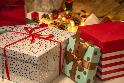 A festive arrangement of wrapped Christmas gift boxes in various sizes and patterns, each tied with decorative ribbons. Warm yellow lights, pinecones, and miniature houses in the background.