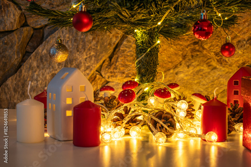 A decorated Christmas tree sparkles with ornaments, tinsel, and fairy lights. Surrounding the tree are wrapped gift boxes, pinecones, miniature houses, and additional red and white candles.