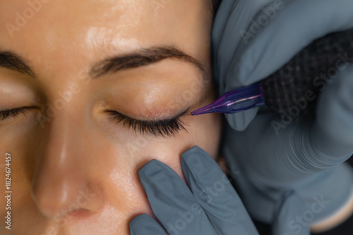 Close up of a cosmetologist performing eyelid permanent makeup, precision eyeliner tattoo procedure with sterile tools, enhancing lash line and eye beauty in a professional studio.