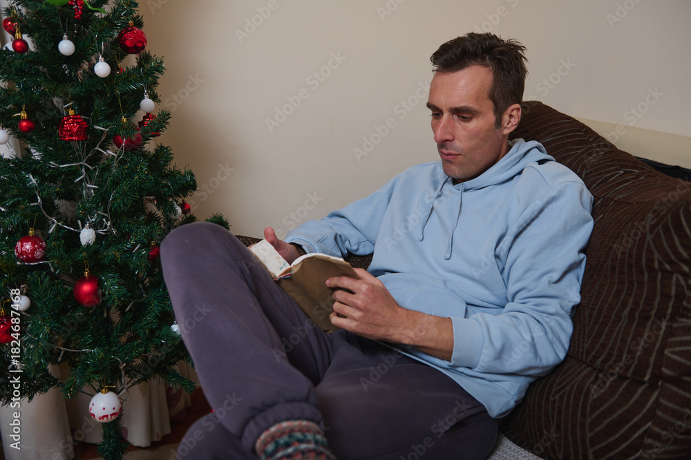 Fototapeta premium Relaxed Man Reading a Book on the Couch Next to the Christmas Tree at Home