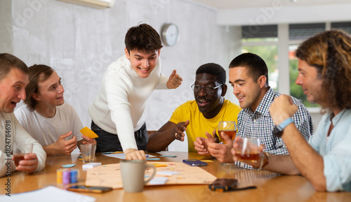 Tableau sur toile Excited man standing on foot playing board game with interested guys sitting aro