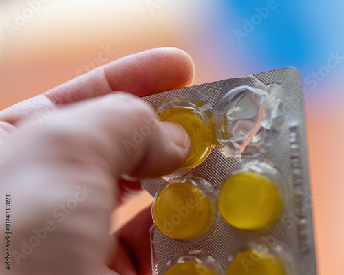 Close-up of a hand pressing out a yellow round lozenge from a silver blister pack against a blurred background.