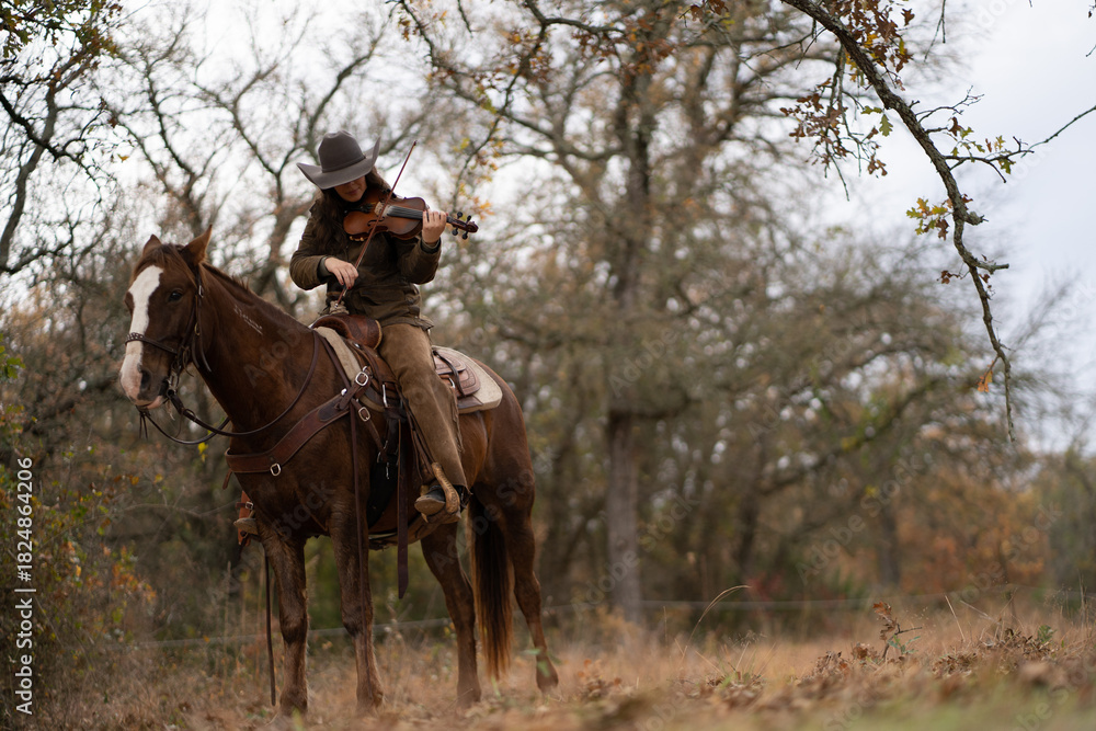 Obraz premium Cowgirl Woman Wearing Cowboy Hat Looking Down and Playing Fiddle Violin While Sitting On Brown and White Horse In Front of Trees and Cloudy Sky