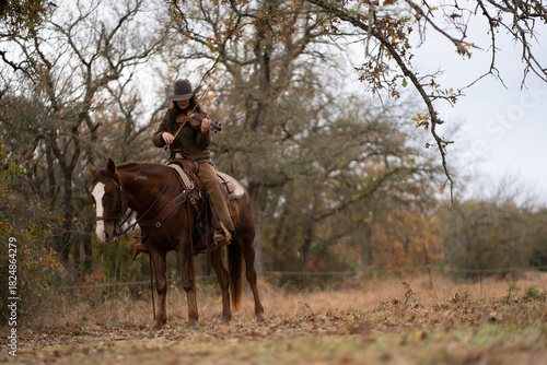Cowgirl Woman Wearing Cowboy Hat Looking Down and Playing Fiddle Violin While Sitting On Brown and White Horse In Front of Trees and Cloudy Sky
