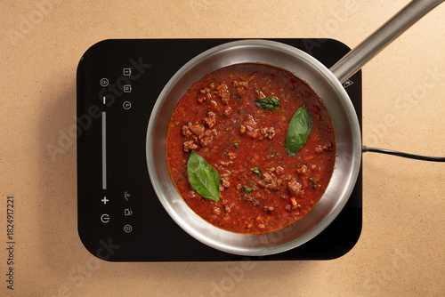 A rich meat sauce bubbles gently in a stainless steel pan with fresh basil leaves on top. This scene depicts a modern kitchen with a sleek induction cooktop ready for cooking.