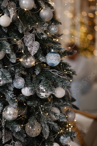Silver and blue Christmas ornaments hanging on a decorated tree with warm lights in a cozy room. Closeup festive scene suitable for holiday, winter, celebration, decoration concepts. 