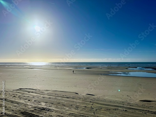 Seascape of the Fort-Mahon and Quend shoreline in Hauts-de-France, expansive sand flats with tidal pools, sparkling backlit waves and a lone silhouette, peaceful winter afternoon under cloudless sky