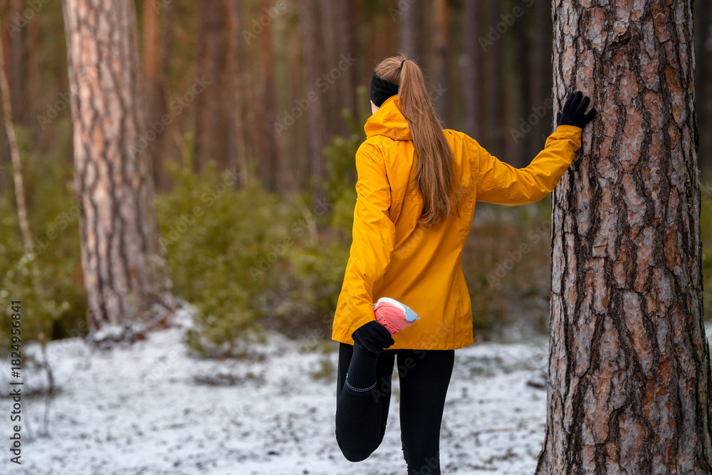 Naklejka premium Woman stretching in a snowy forest while holding a pine tree, warming up before a winter run, focused and calm in cold natural surroundings.