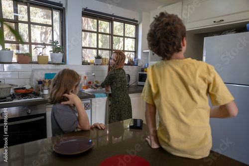 A woman with bright dreadlocks is focused on washing dishes in the kitchen sink, while her two children wait nearby at the counter, reflecting an attentive and diligent mood.