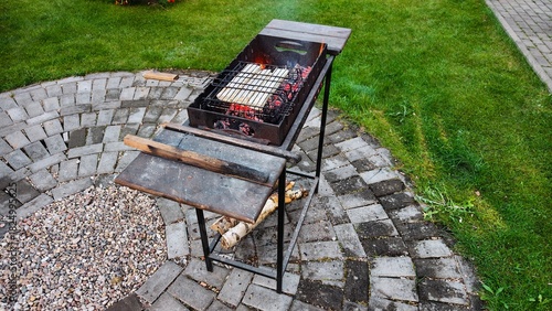 On a tiled area near a grassy lawn there is a metal brazier in which coals are smoldering, and lavash with cheese is being cooked on the grill. There are firewood nearby. Autumn evening twilight
