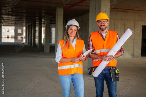 Two construction workers, a man and a woman, wearing safety vests and helmets, stand in the unfinished building, holding blueprints and a tablet, overseeing the progress of their project