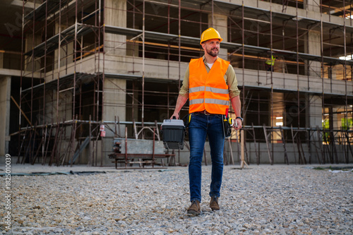 Construction worker in a safety vest and hardhat walking along gravel at a building site, carrying a toolbox filled with essential tools for the job