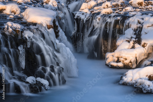 View of cascading waterfalls flow amidst ice-covered rocks, creating a serene winter landscape bathed in soft light, Hlauptunga, Blaskogabyggd, Iceland.